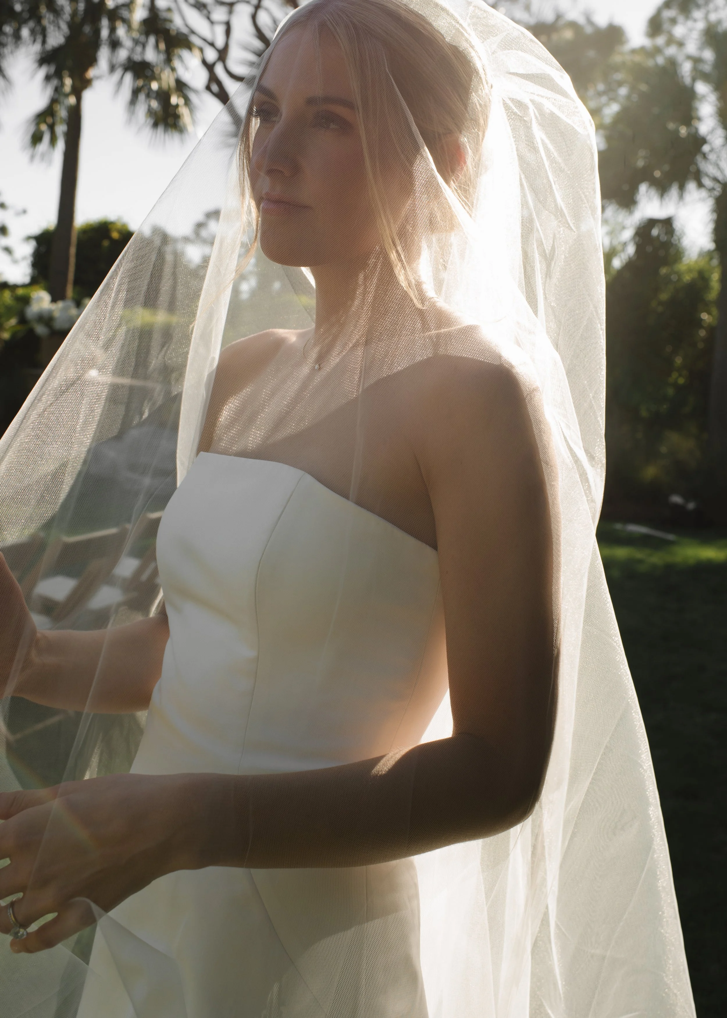 A bride in a strapless wedding dress wearing a veil standing outside with sunlight illuminating her face and shoulder.