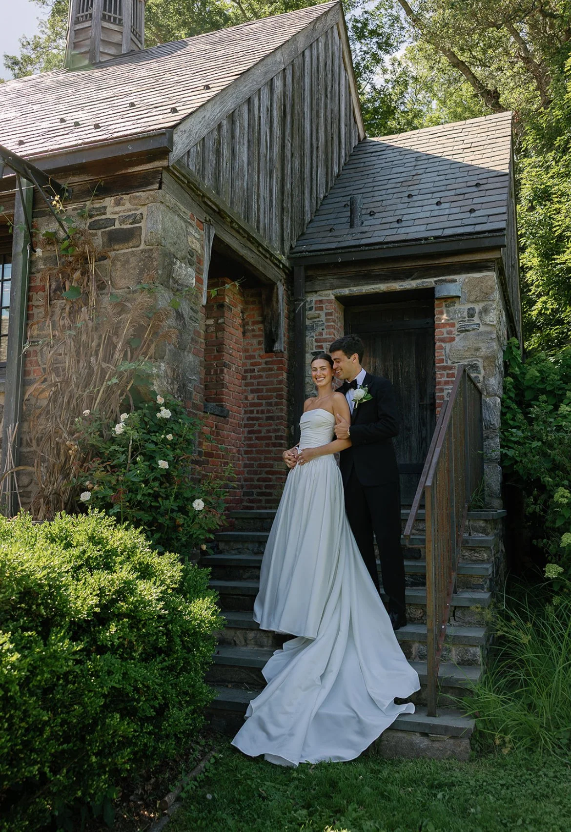 A bride and groom standing together on stone steps in front of a rustic brick and wood building, smiling and enjoying their wedding day amid lush green trees and bushes.