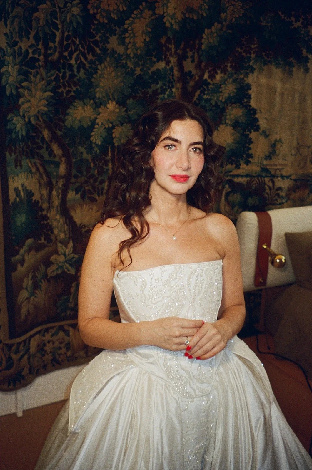 A woman with curly dark hair, wearing a white wedding dress with intricate beadwork, standing indoors with a nature-themed tapestry in the background.