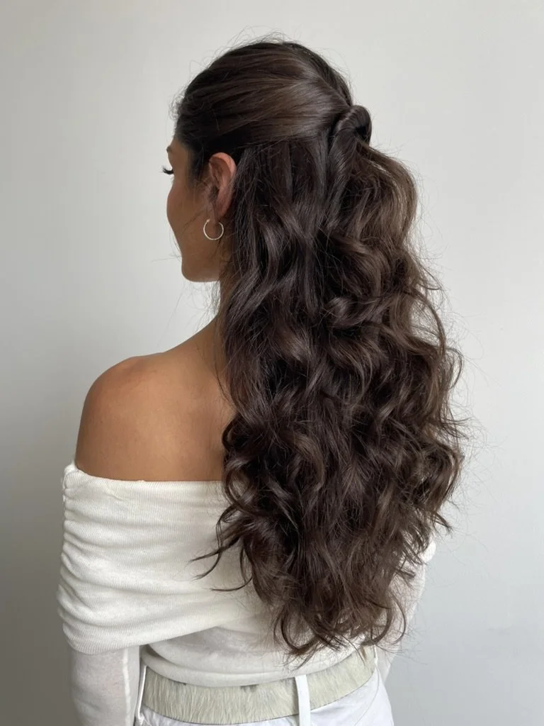 Side view of a woman with long, wavy brown hair tied in a half-up hairstyle, wearing a white off-the-shoulder top and hoop earrings, standing against a plain white background.