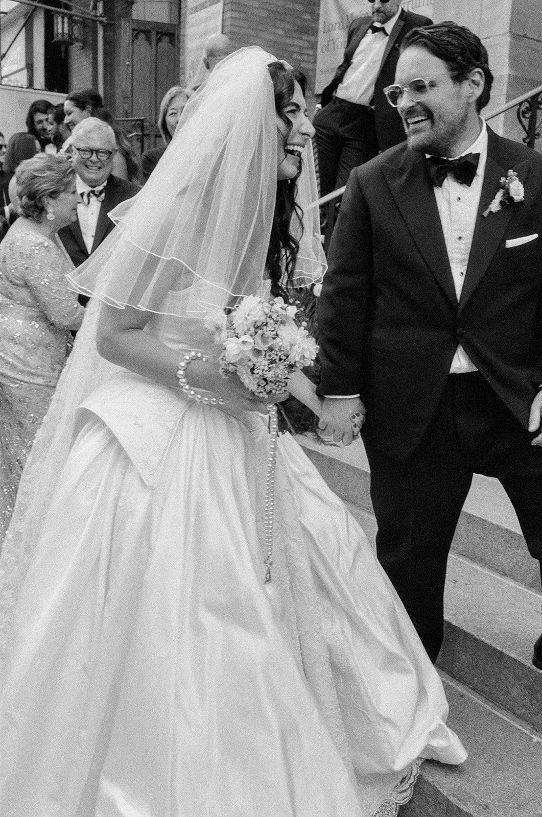 Black and white photo of a wedding, featuring a bride in a wedding gown and veil holding hands with a groom in a tuxedo, surrounded by smiling guests.