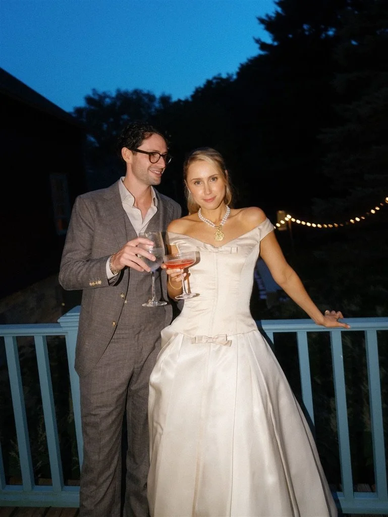 A man in a gray suit and glasses, and a woman in a white wedding dress, standing outdoors on a deck at night, holding glasses of wine, with string lights in the background.