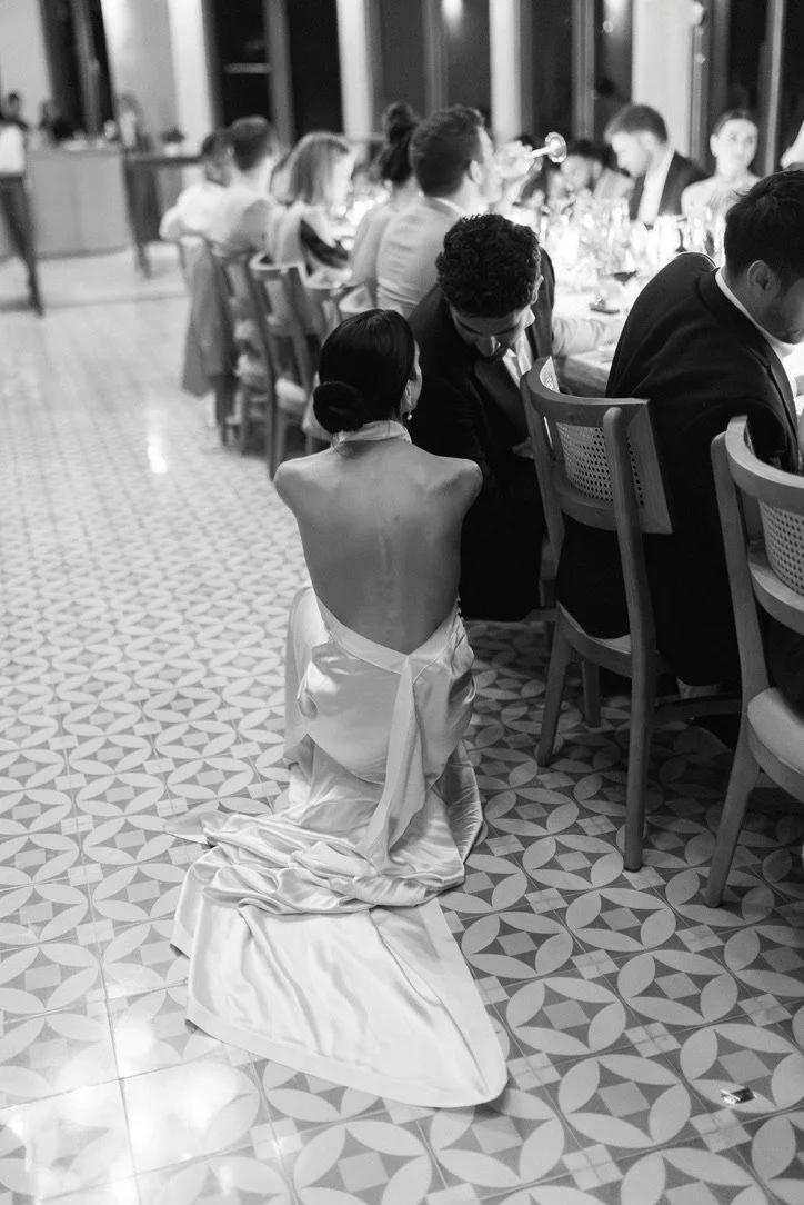A woman in a wedding dress kneeling on the patterned floor, leaning towards a man at a formal dinner event.