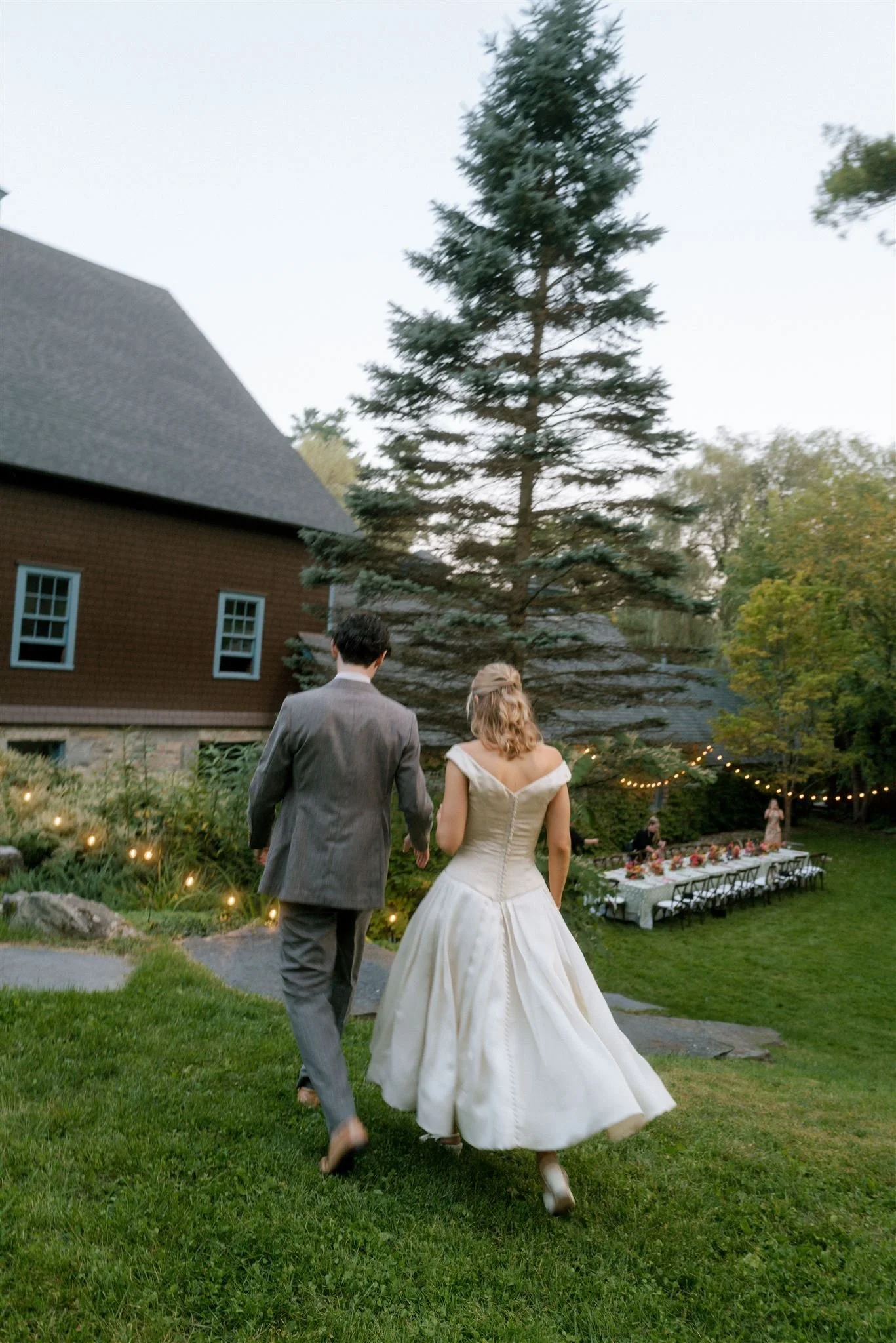 A bride and groom walking in a garden toward a wedding reception area with tables, chairs, and string lights in the background.