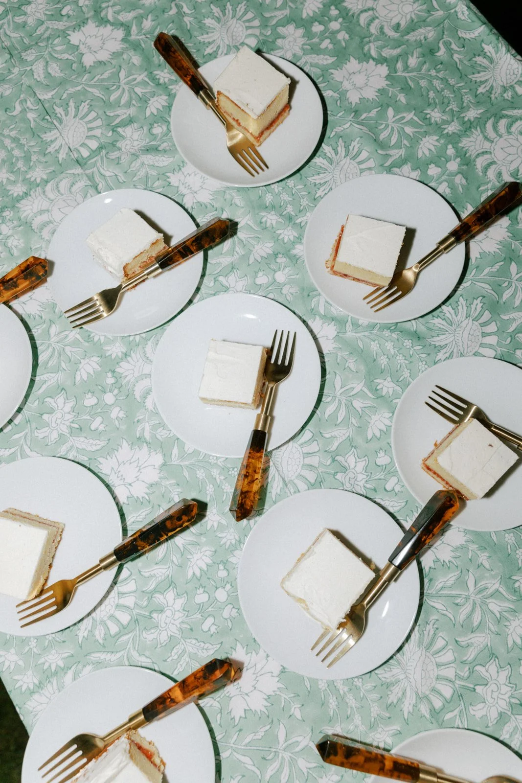 Multiple white plates each with a slice of cake and a fork on a patterned tablecloth.