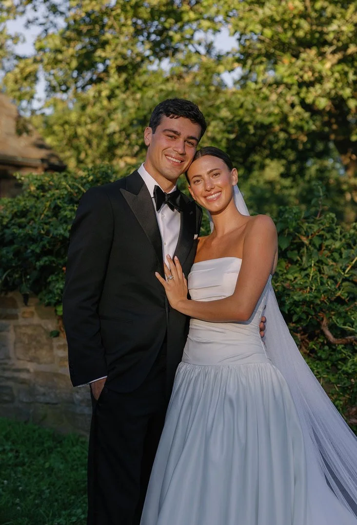 A happy couple in wedding attire outdoors, with trees and bushes in the background. The groom wears a black tuxedo with a black bow tie, and the bride wears a white strapless wedding gown with a veil.