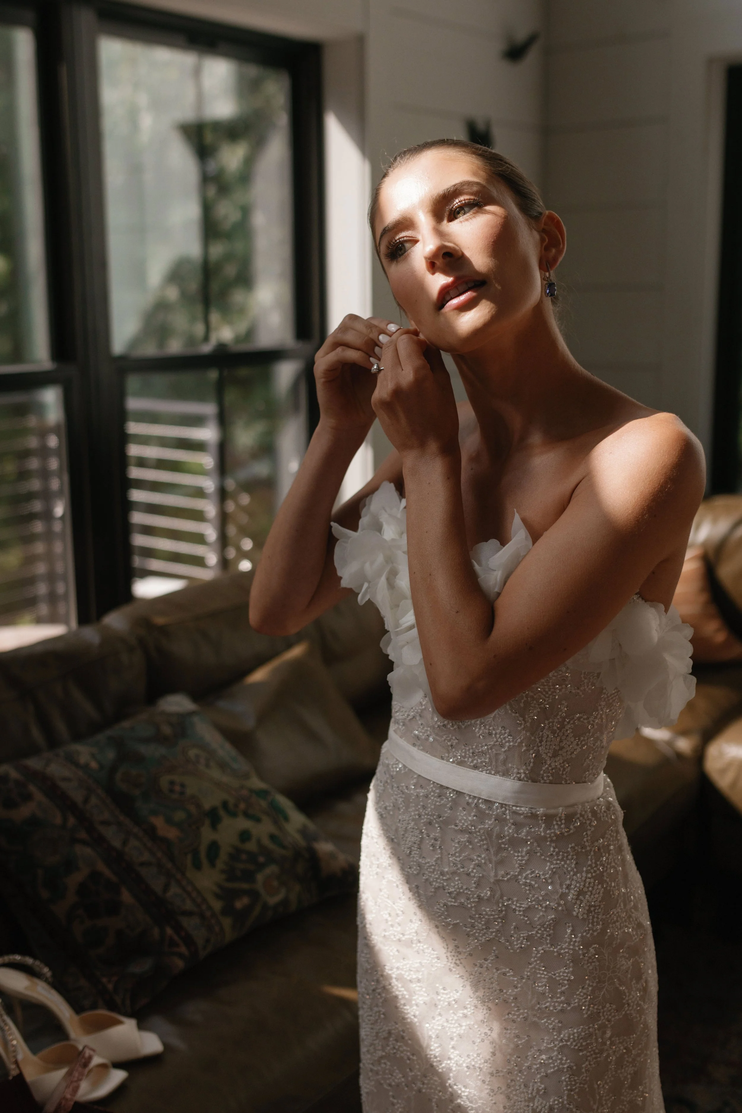 A bride in a white wedding gown adjusting her earring in a sunlit room with large windows and a brown sofa in the background.