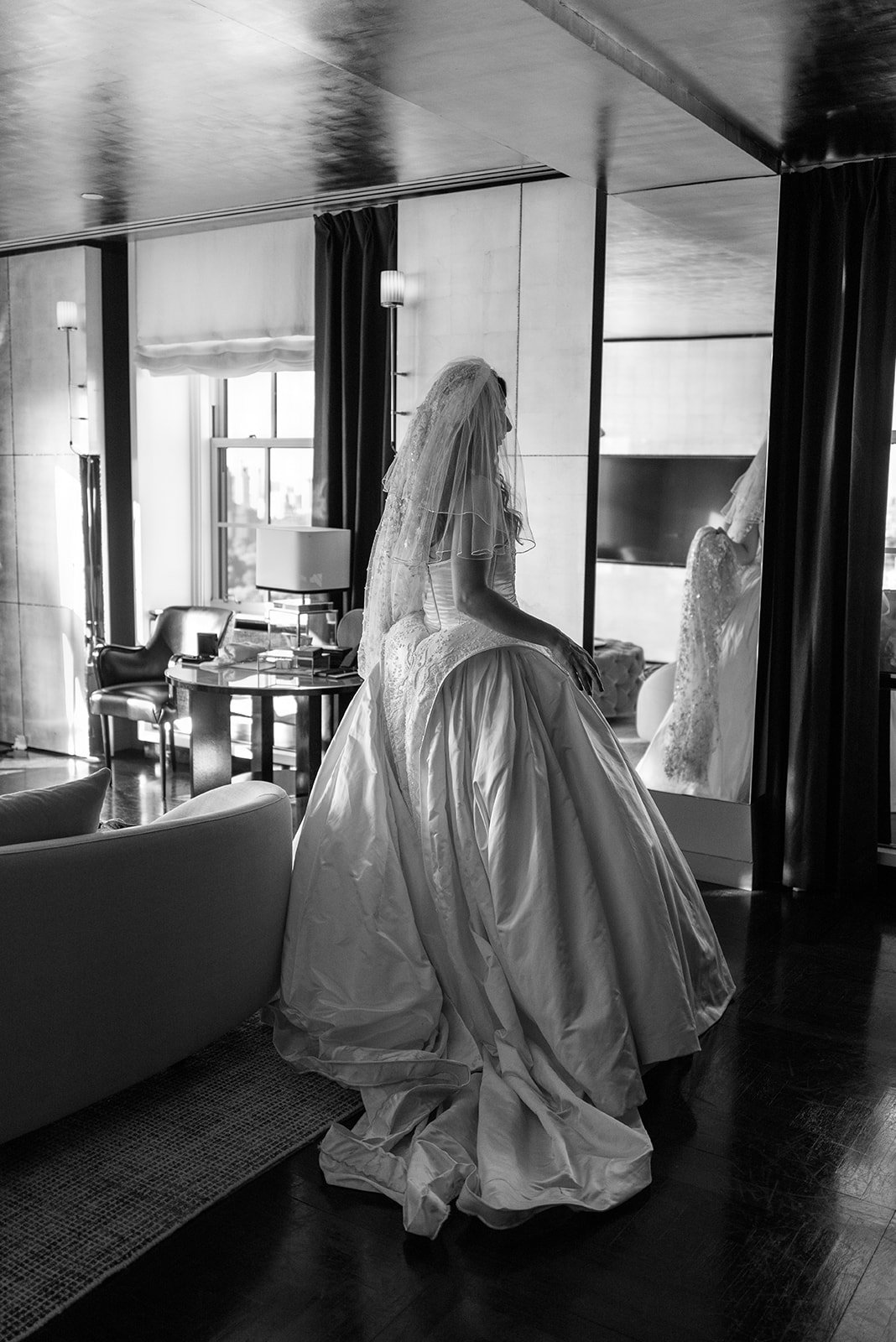A bride in a wedding gown sitting on a chair, looking at her reflection in a mirror in a hotel room.