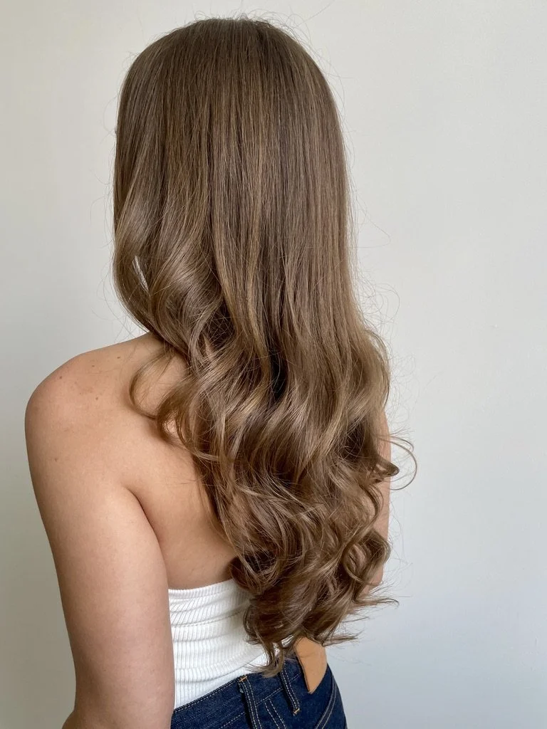A woman with long, wavy, light brown hair wearing a white strapless top and blue jeans, standing against a plain white wall.