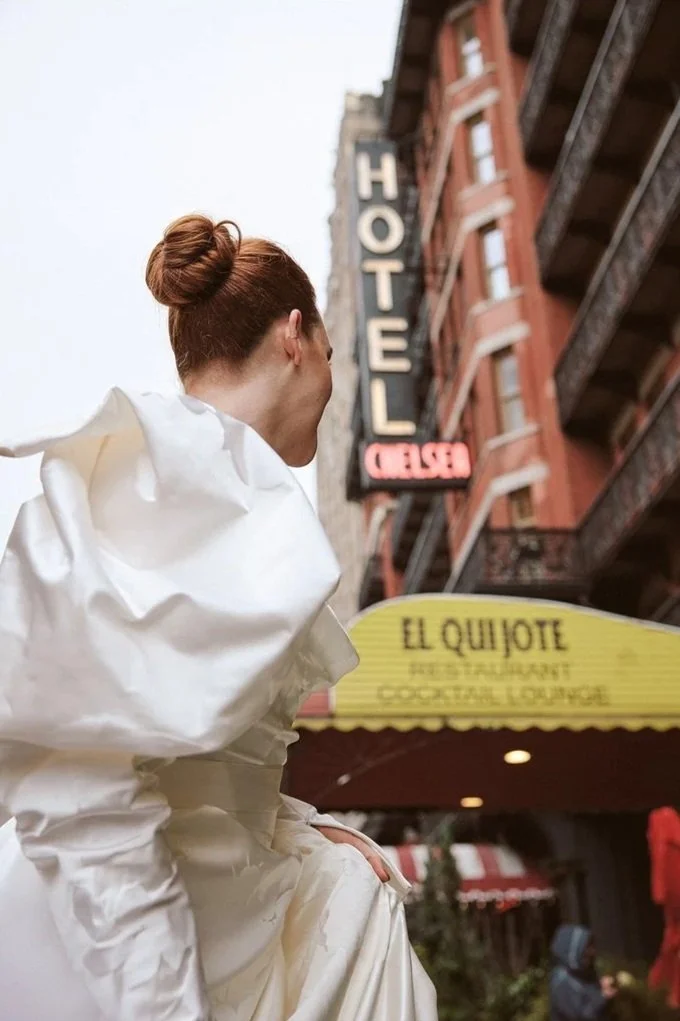 A woman sitting on a ledge outside a building with a 'HOTEL' sign, wearing a white garment with her hair in a bun, looking away from the camera.