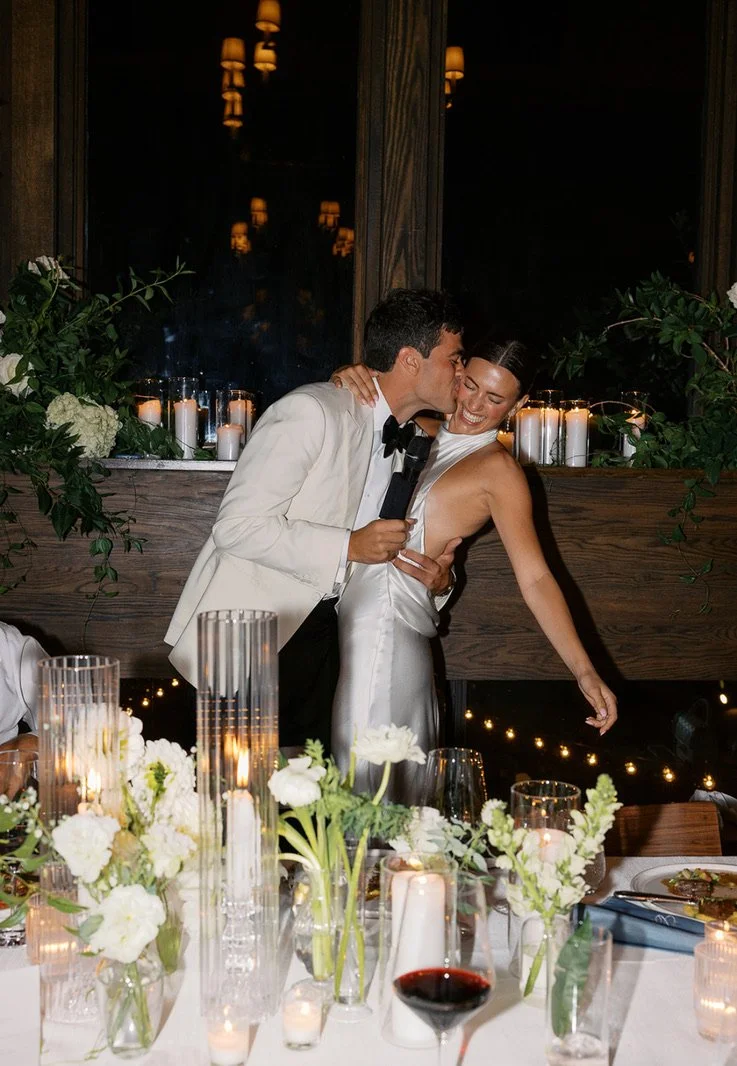 A couple, dressed in wedding attire, sharing a kiss at their wedding reception. The groom is in a white tuxedo, and the bride is in a white satin gown. There are candles and floral arrangements on the table in front of them.