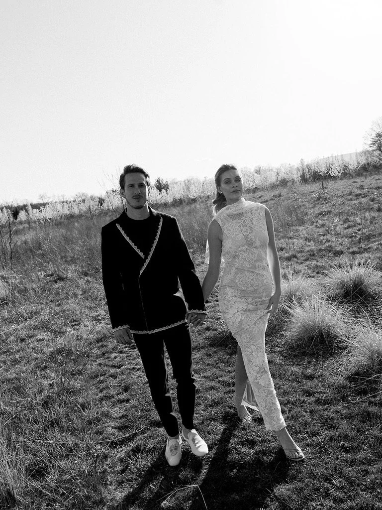 A man and woman holding hands walking outdoors in a grassy field, dressed in fashionable clothing, with a bright sky above.