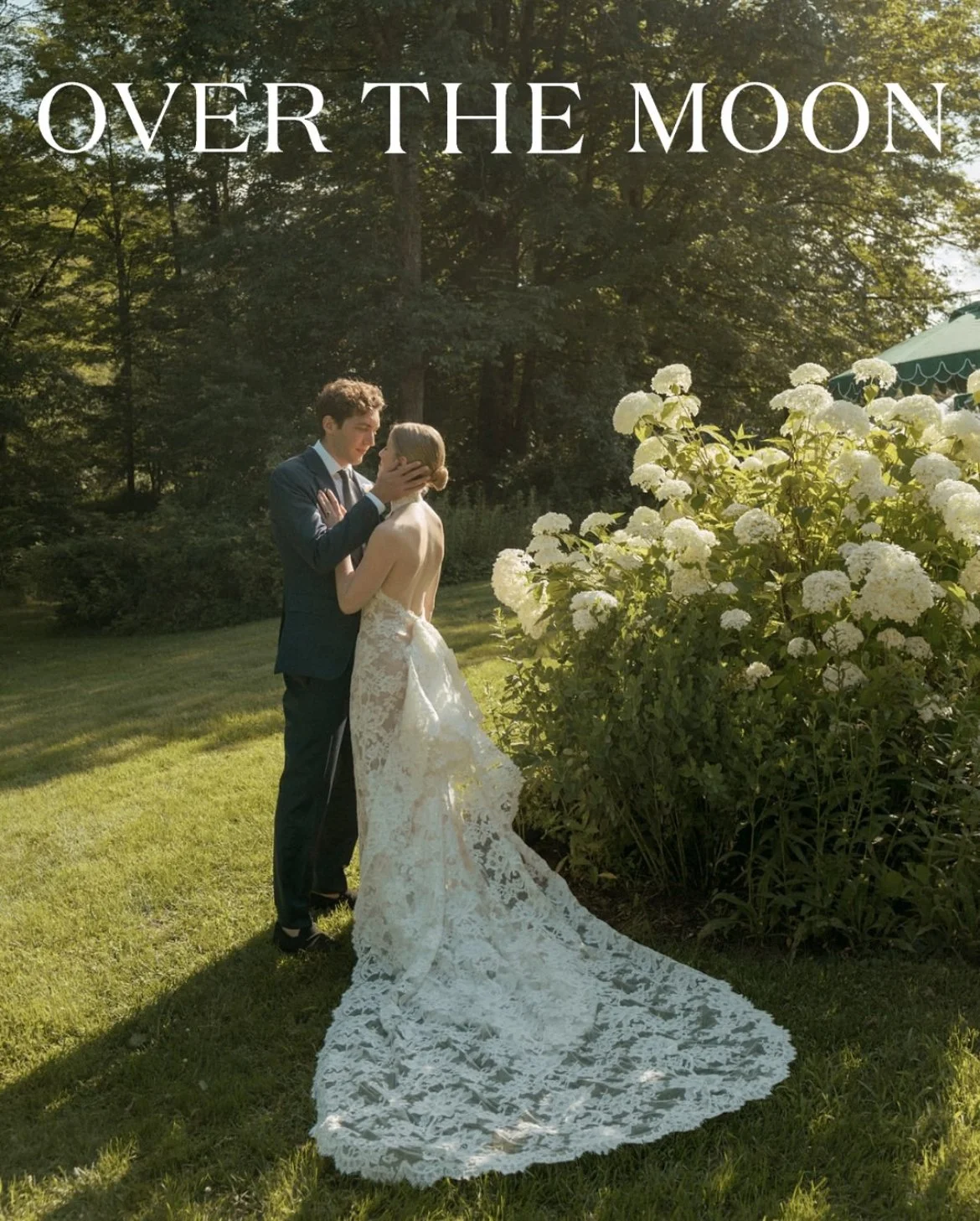 A couple in wedding attire sharing a kiss outdoors during daytime, with lush green grass, trees, and white flowers in the background, and the words 'Over the Moon' written at the top.