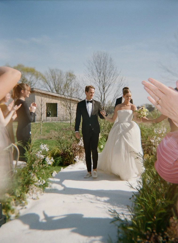 Bride and groom walking down an outdoor aisle, surrounded by guests clapping, with a building and bare trees in the background on a sunny day.