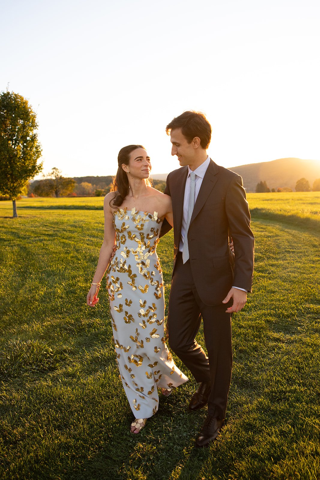 A woman and man walking together on a grassy field at sunset. The woman is wearing a white dress with gold butterfly embellishments, and the man is dressed in a dark suit with a light-colored tie. They are looking at each other and smiling.