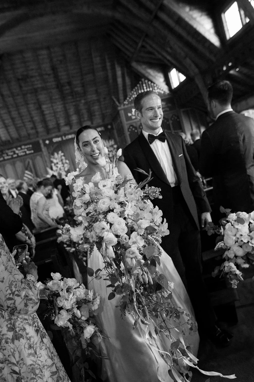 A black and white photo of a bride and groom during their wedding ceremony inside a church. The bride is holding a large bouquet of flowers, and both are smiling. The church has a high, wooden ceiling and guests are visible in the background.