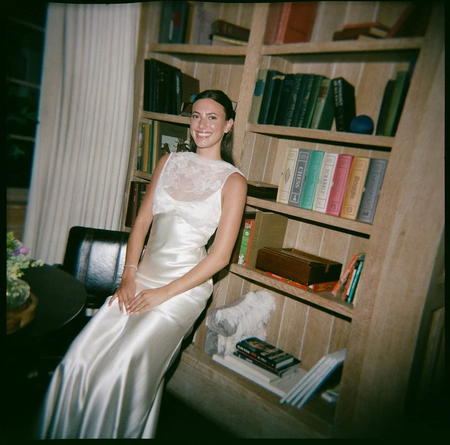 A woman in a white satin dress sitting on a black chair in a room with wooden bookshelves filled with books.