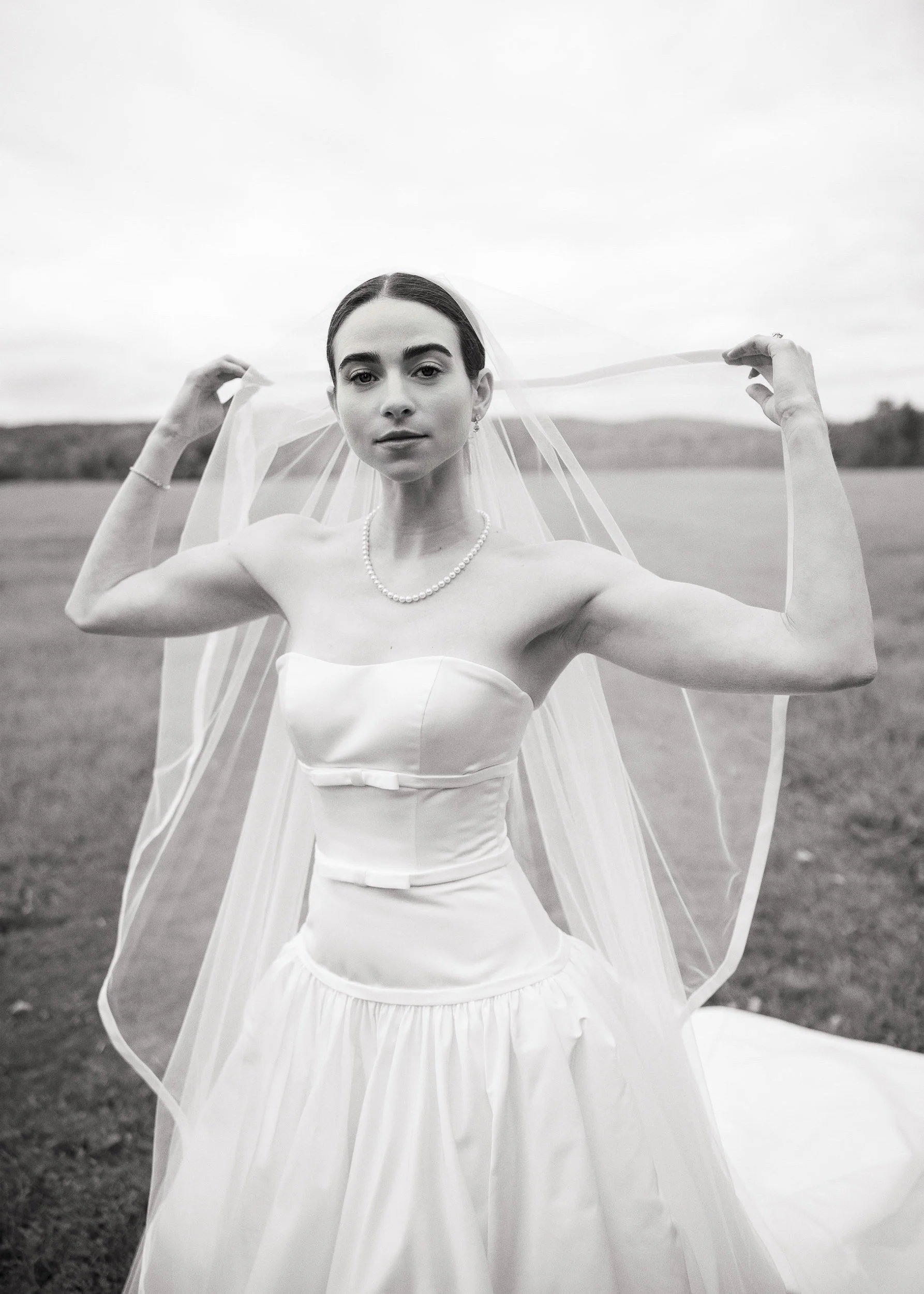 A black and white photo of a woman in a wedding dress, standing outdoors in a field, looking directly at the camera, holding her veil with both hands.