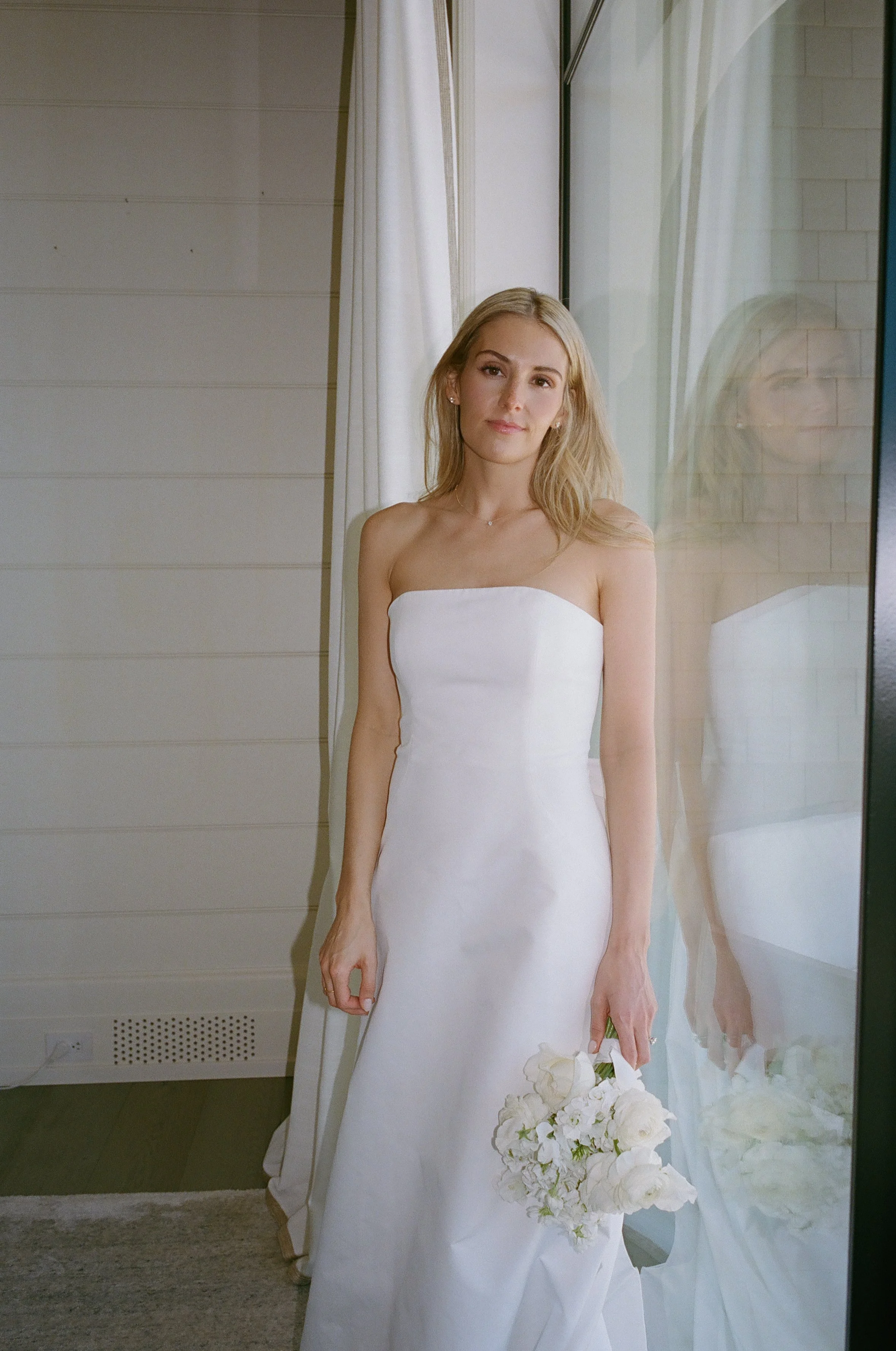 A woman in a strapless white wedding dress holding a bouquet of white flowers standing near a window with white curtains, with her reflection visible in the glass.
