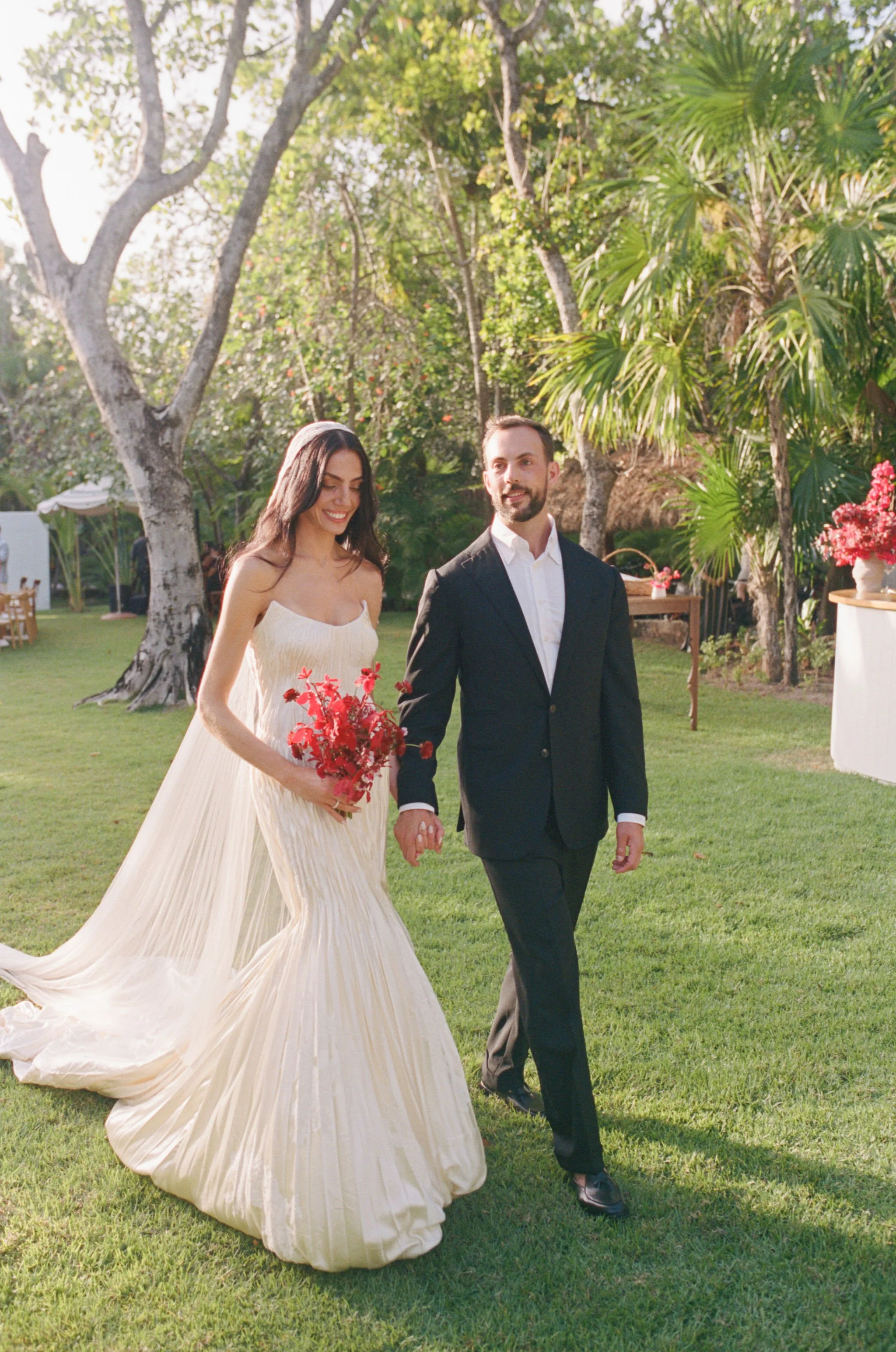 A bride in a flowing white gown holding a bouquet of red flowers, walking hand-in-hand with a groom in a black suit outdoors surrounded by lush green trees and plants.