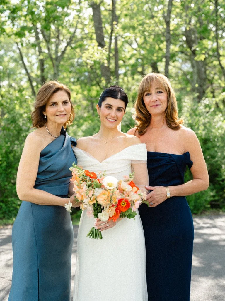 A bride in a white wedding dress holding a colorful bouquet of flowers, standing with two women outdoors in a green wooded area.