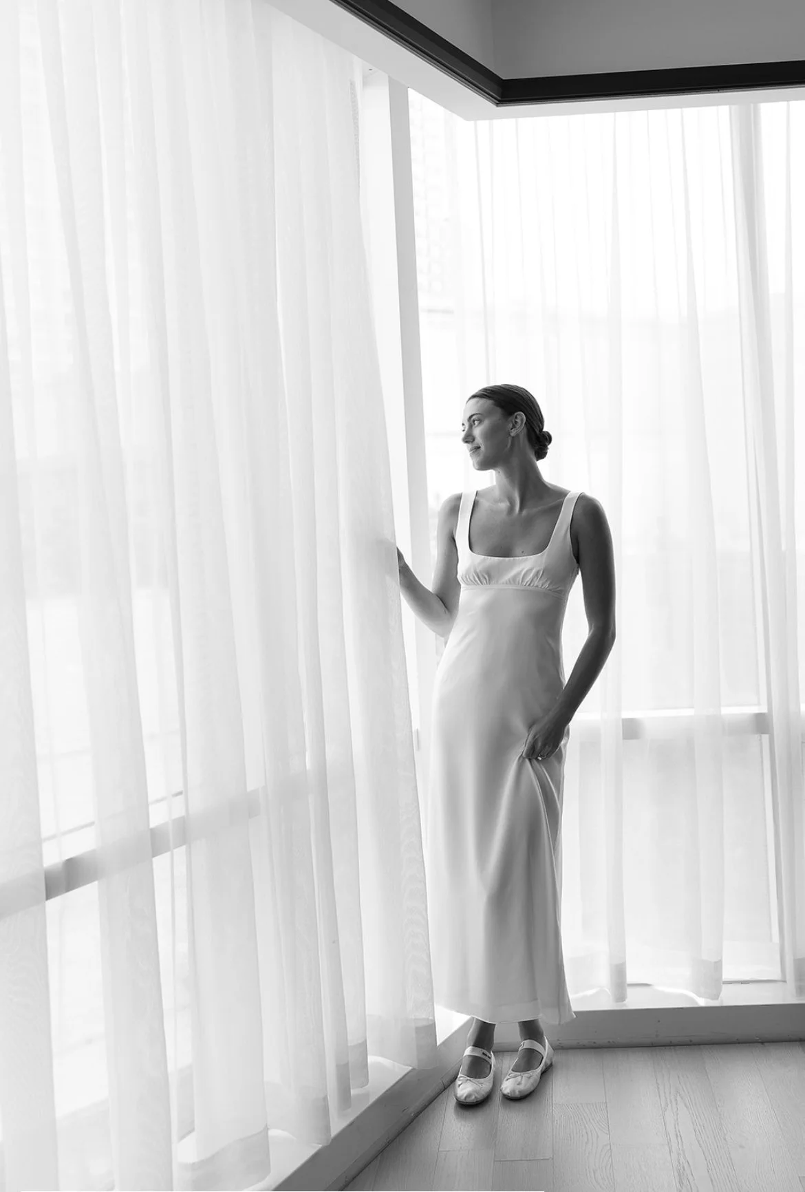 Black and white photo of a woman in a dress by a window with sheer curtains, standing and looking outside.