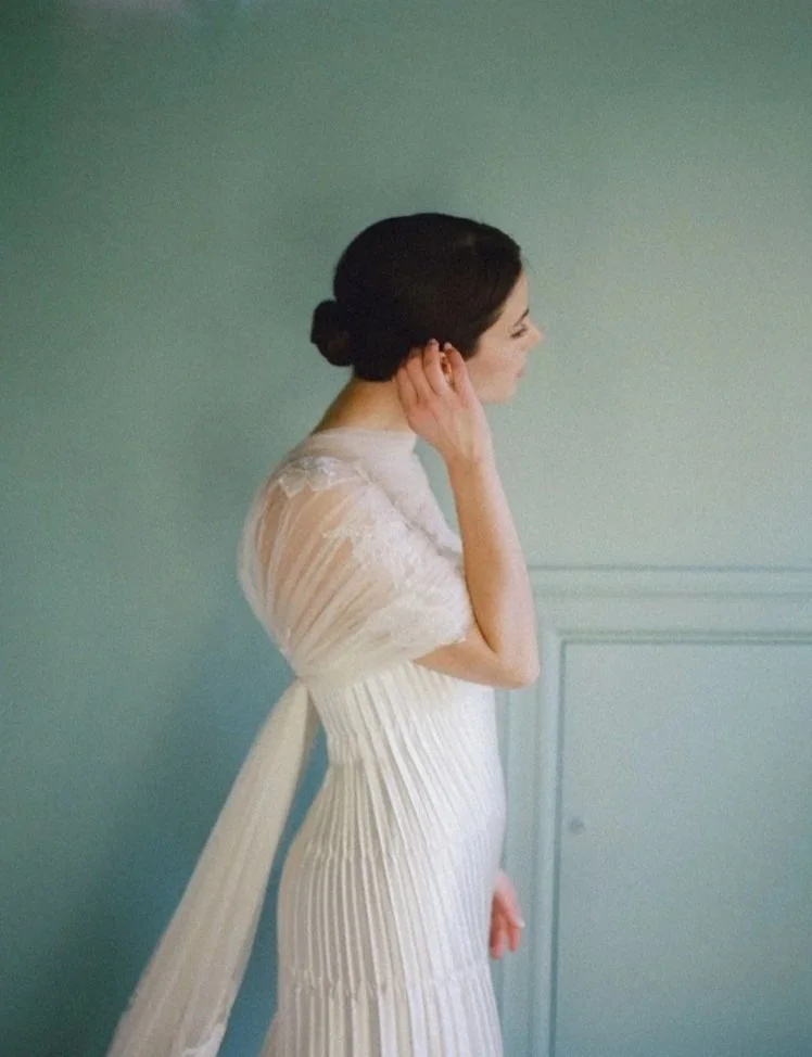 A woman in a white dress with pleats and lace details, with dark hair styled in an elegant bun, standing in profile against a light green wall.
