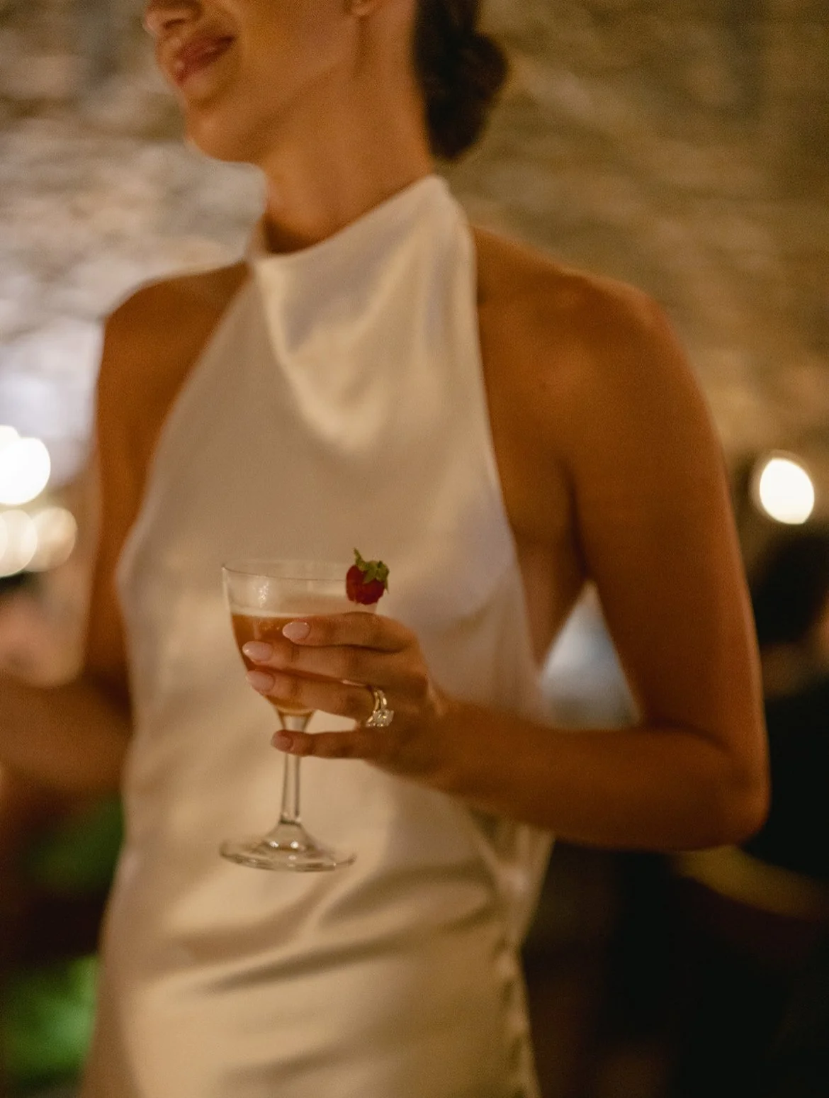 A woman in a white satin halter dress holding a cocktail with a strawberry garnish, in a dimly lit setting.