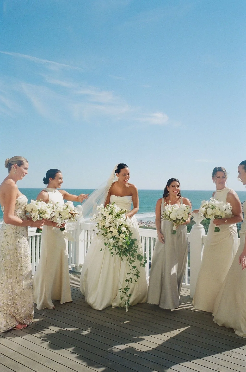 A bride in a strapless white wedding dress and veil, surrounded by five bridesmaids holding white floral bouquets, standing on a beachside balcony with ocean view under a clear blue sky.