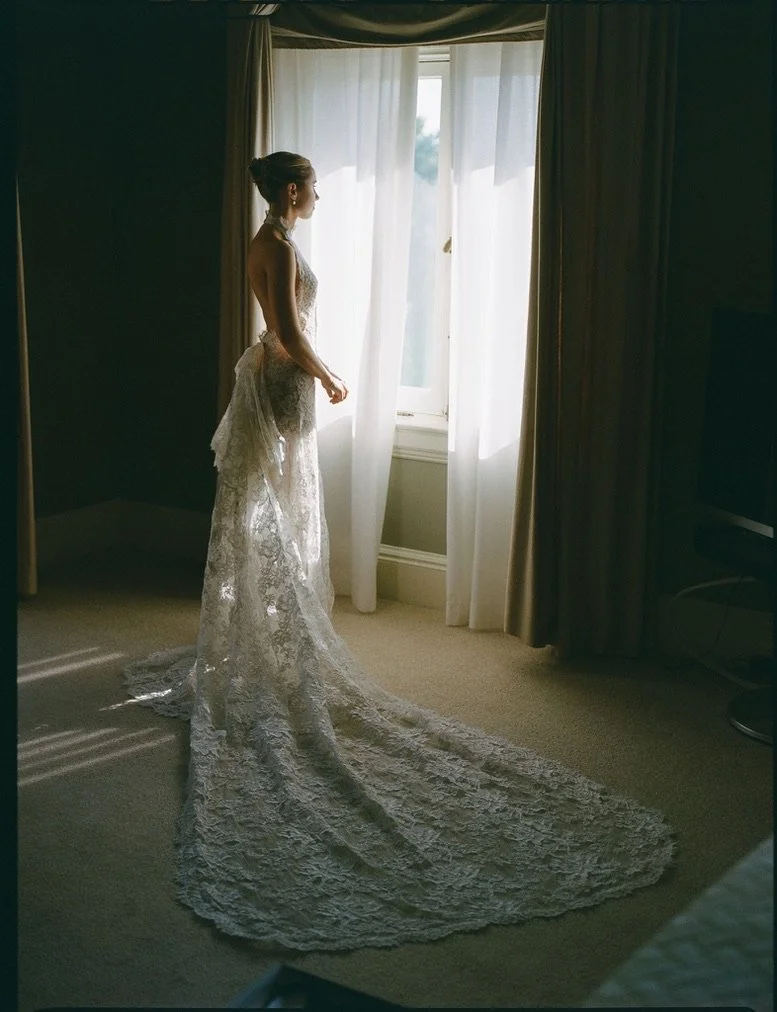 A woman in a white lace wedding dress standing by a window in a room, looking outside.