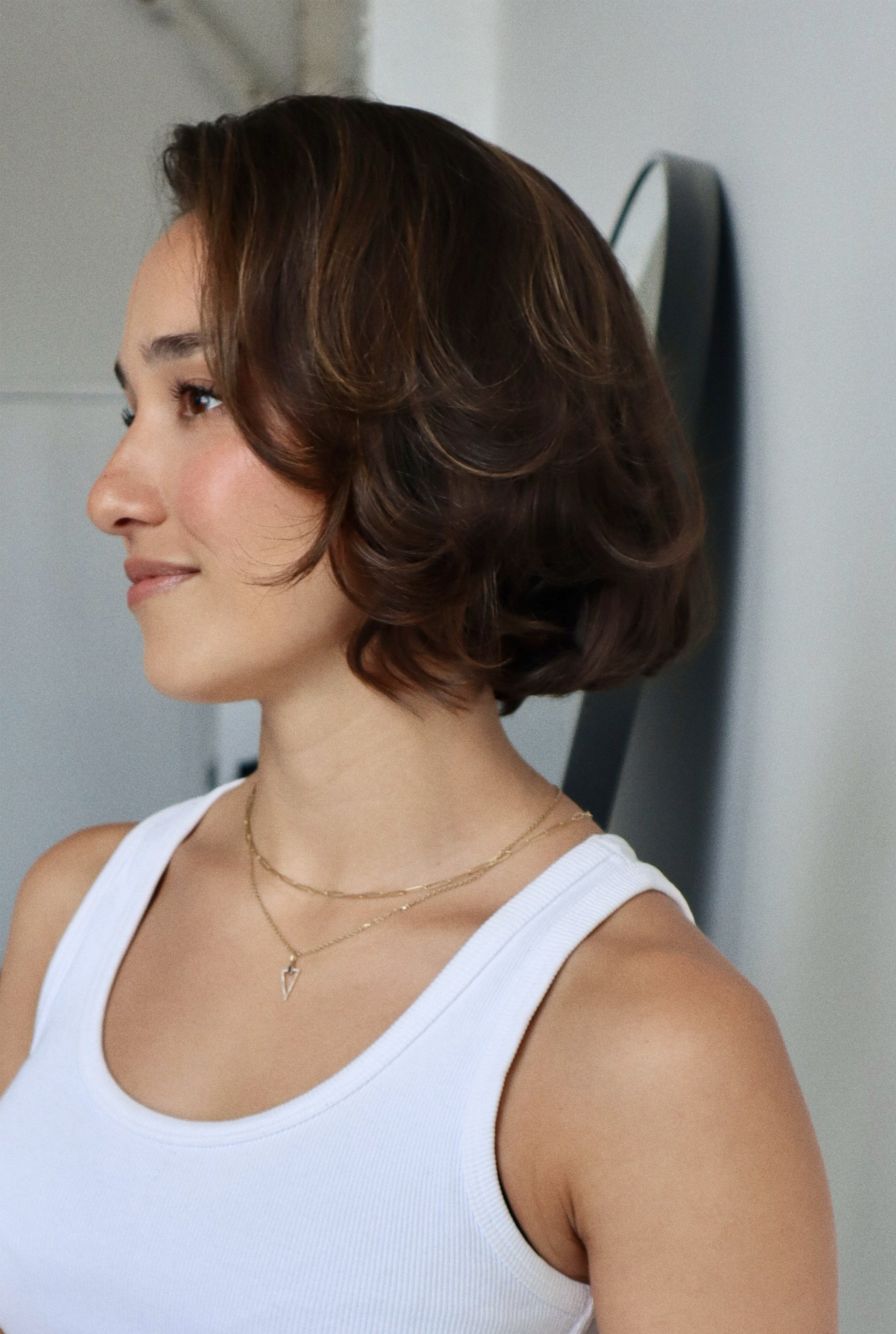 A woman with short, wavy brown hair, wearing a white tank top and layered gold necklaces, looking to her left indoors.