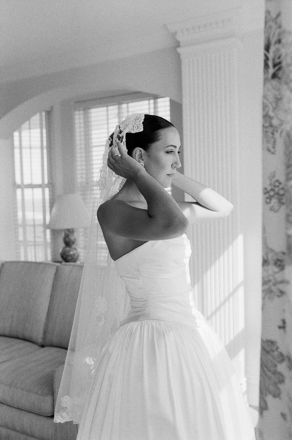 Black and white photo of a bride adjusting her veil indoors near a window with blinds, a patterned wallpaper, and a sofa.
