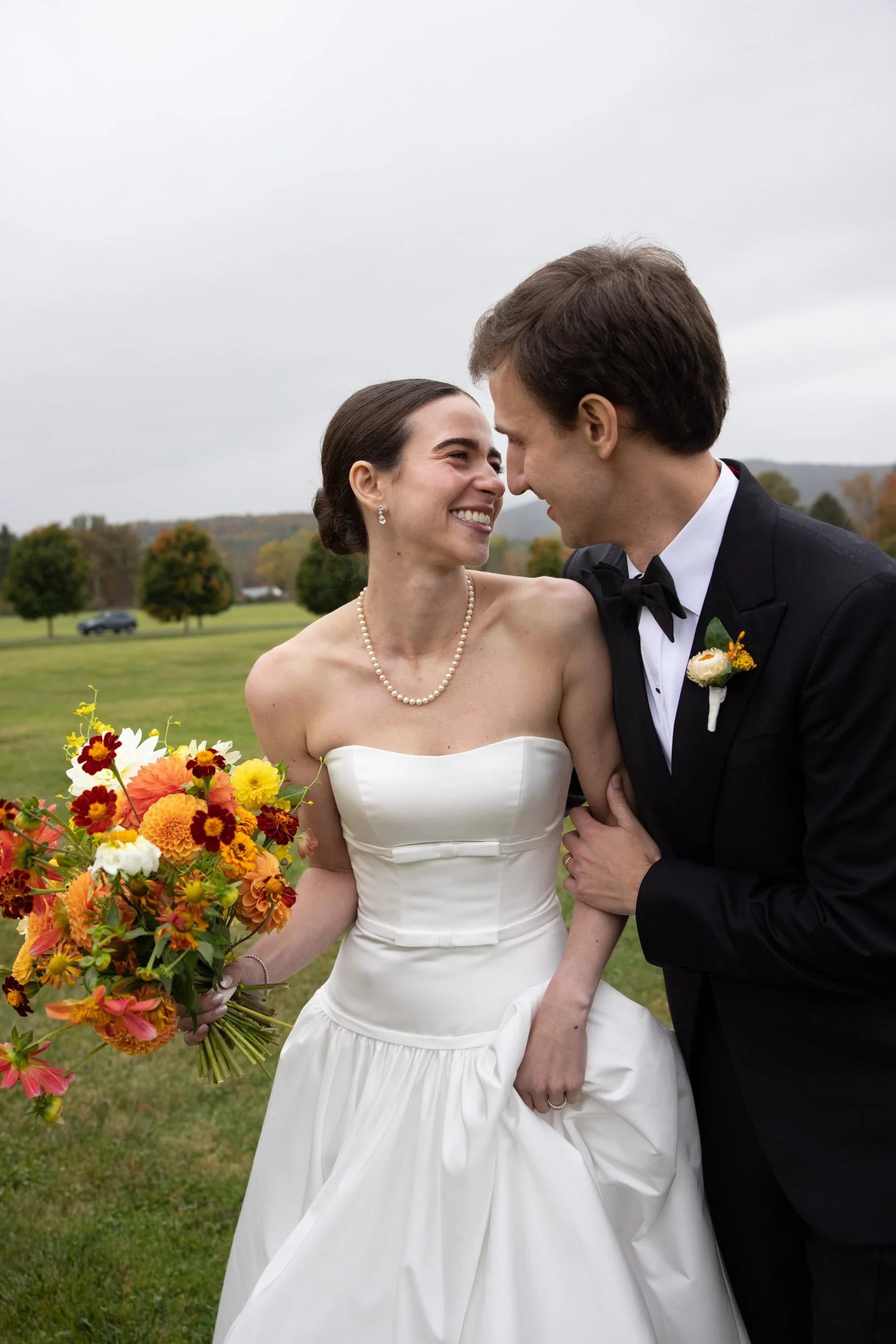 A bride and groom smiling at each other outdoors on their wedding day. The bride wears a white strapless wedding dress, a pearl necklace, and holds a colorful bouquet. The groom wears a black tuxedo with a bowtie and a boutonniere. They are close tog