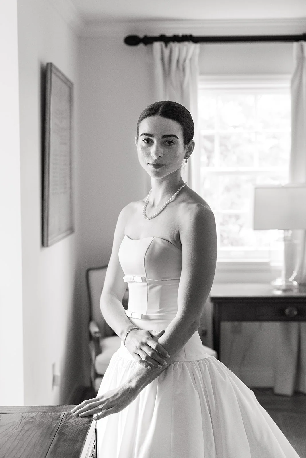 Black and white photo of a young woman in a wedding dress, standing indoors near a wooden table, with a window, curtains, and a lamp in the background.