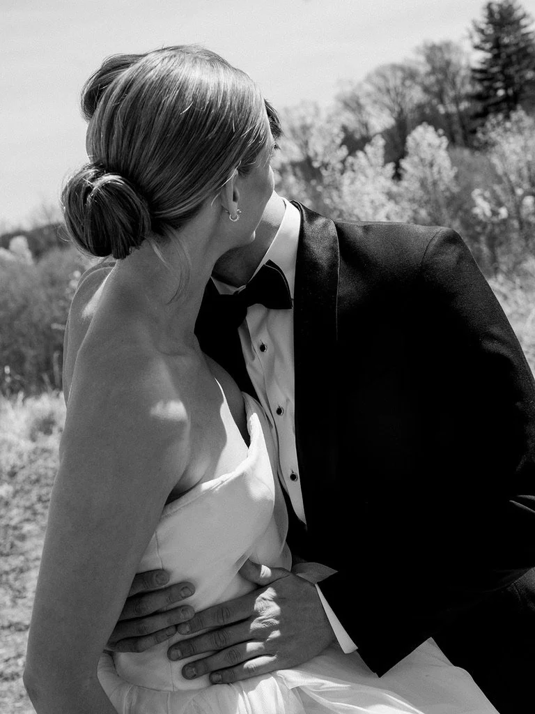 A black and white photo of a bride and groom sharing a kiss outdoors, with trees in the background. The bride is wearing a strapless dress and the groom is in a tuxedo with a bow tie.