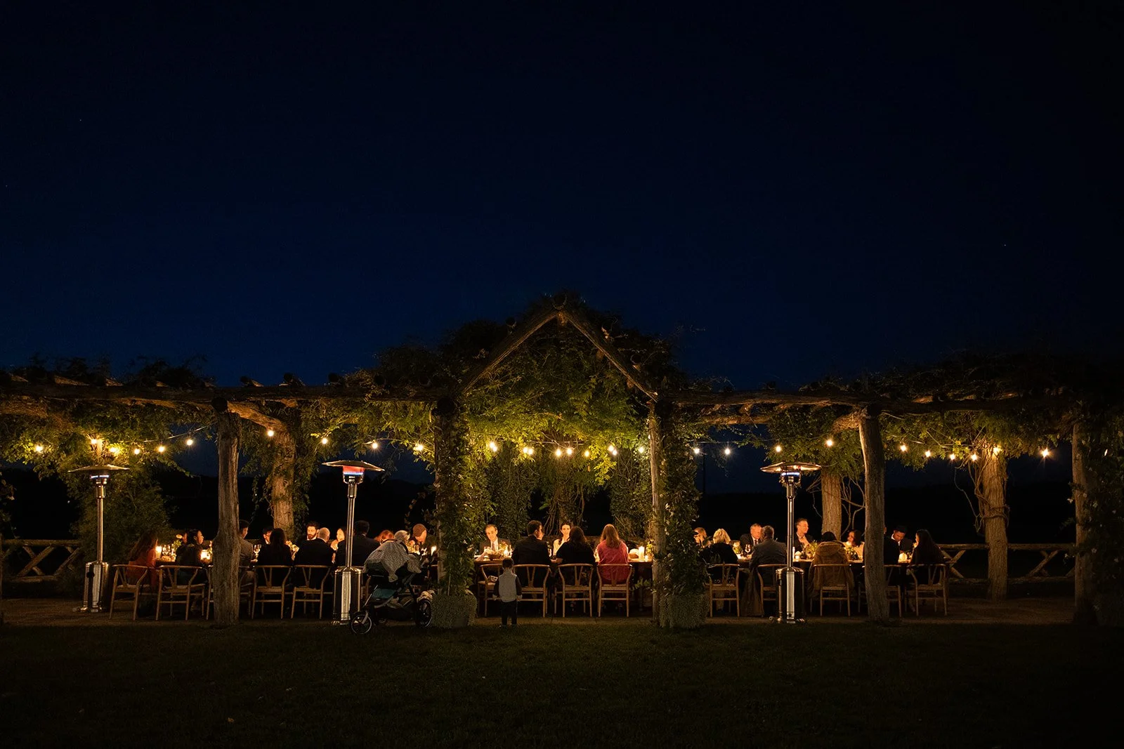 Nighttime outdoor dinner gathering under a wooden pergola decorated with string lights, with guests seated at long tables, outdoors with heaters and a dark sky