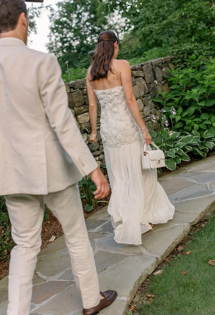 A woman in a white dress holding a white handbag walks along a stone path, with a man in a beige suit walking nearby, outdoors with greenery and a stone wall.