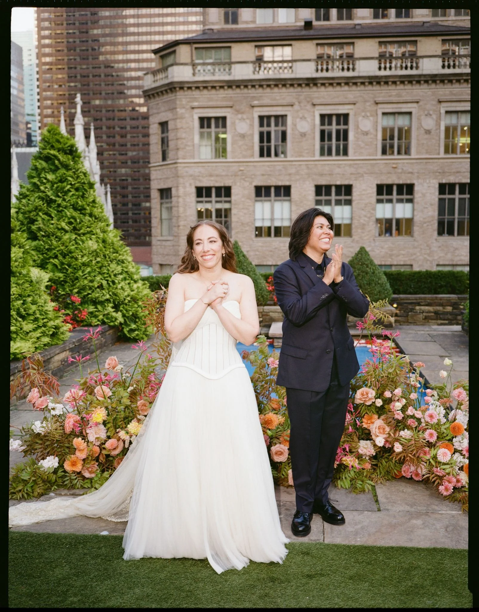 Two women standing outdoors on a paved area with colorful flowers, greenery, and urban buildings in the background. One woman in a white dress is smiling and holding her hands together, and the other woman in a dark suit is also smiling with her hands clasped.