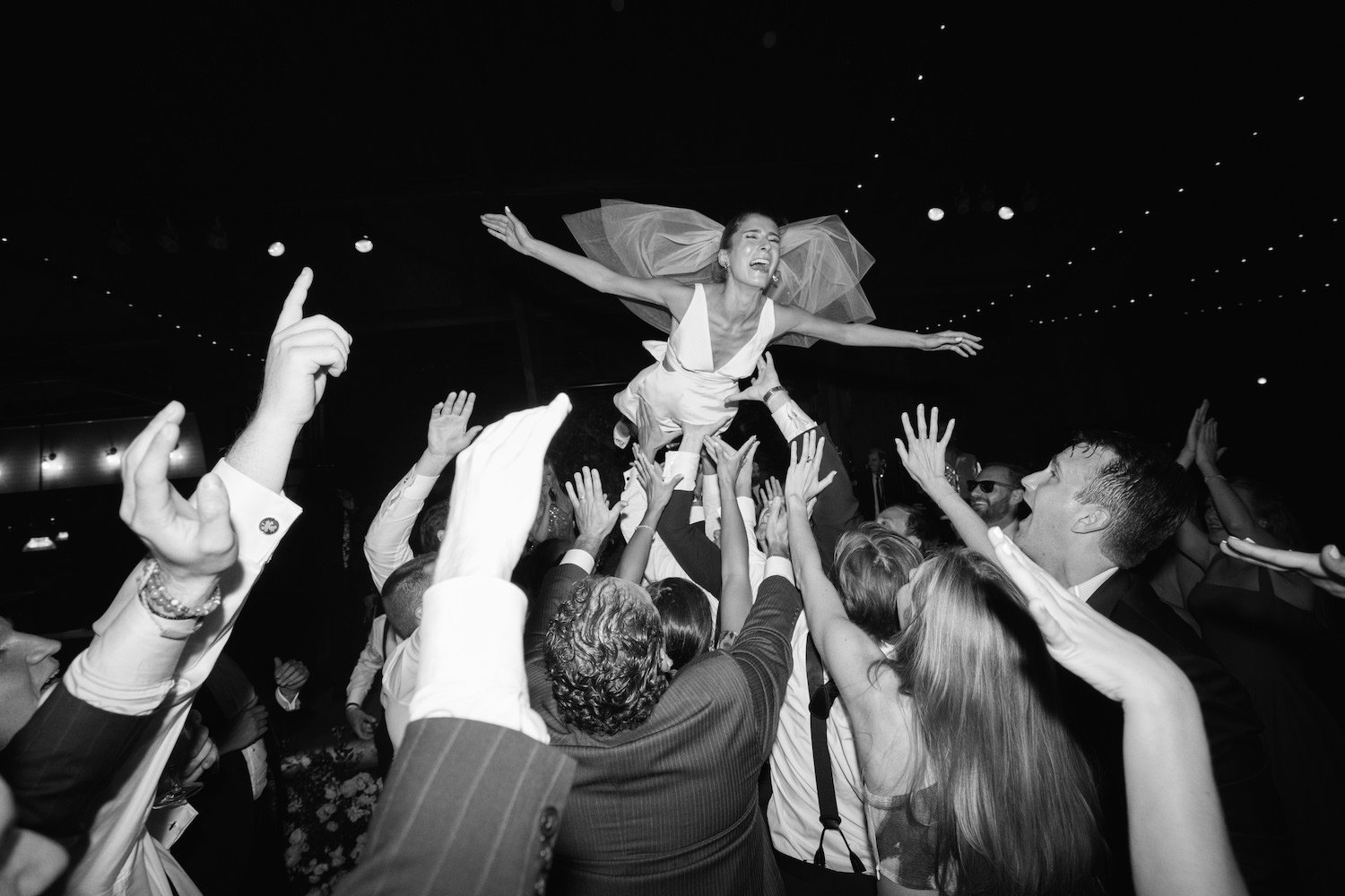 A woman in a wedding dress being lifted into the air by a crowd of people at a celebration, with many people reaching out to support her.