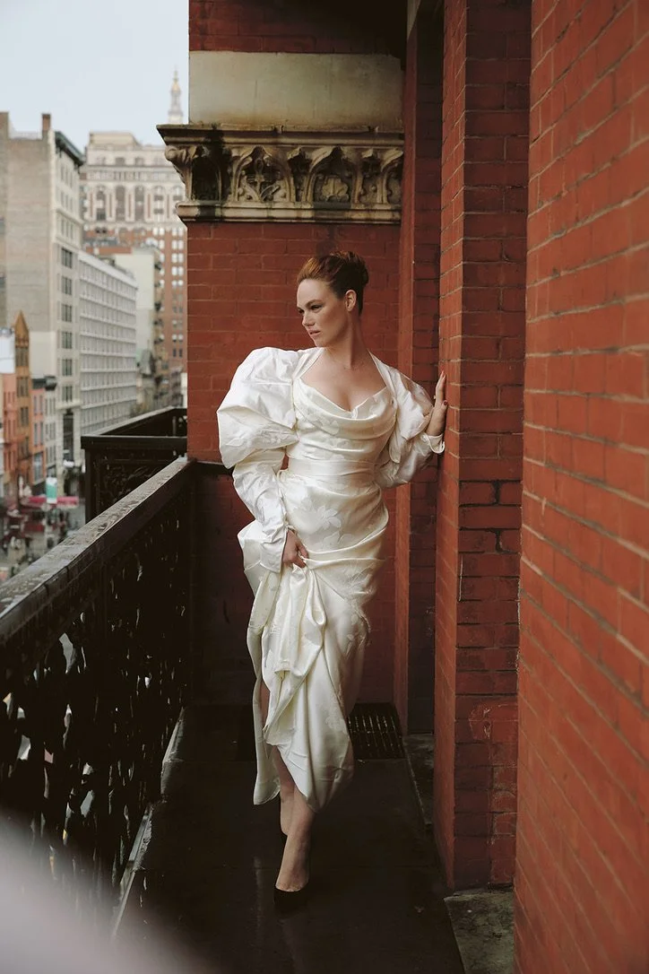 A woman in an elegant, cream-colored satin dress standing on a city rooftop balcony. She is leaning against a brick wall with her left hand and holding her dress with her right, looking contemplative. The balcony has a decorative black metal railing, and city buildings are visible in the background.
