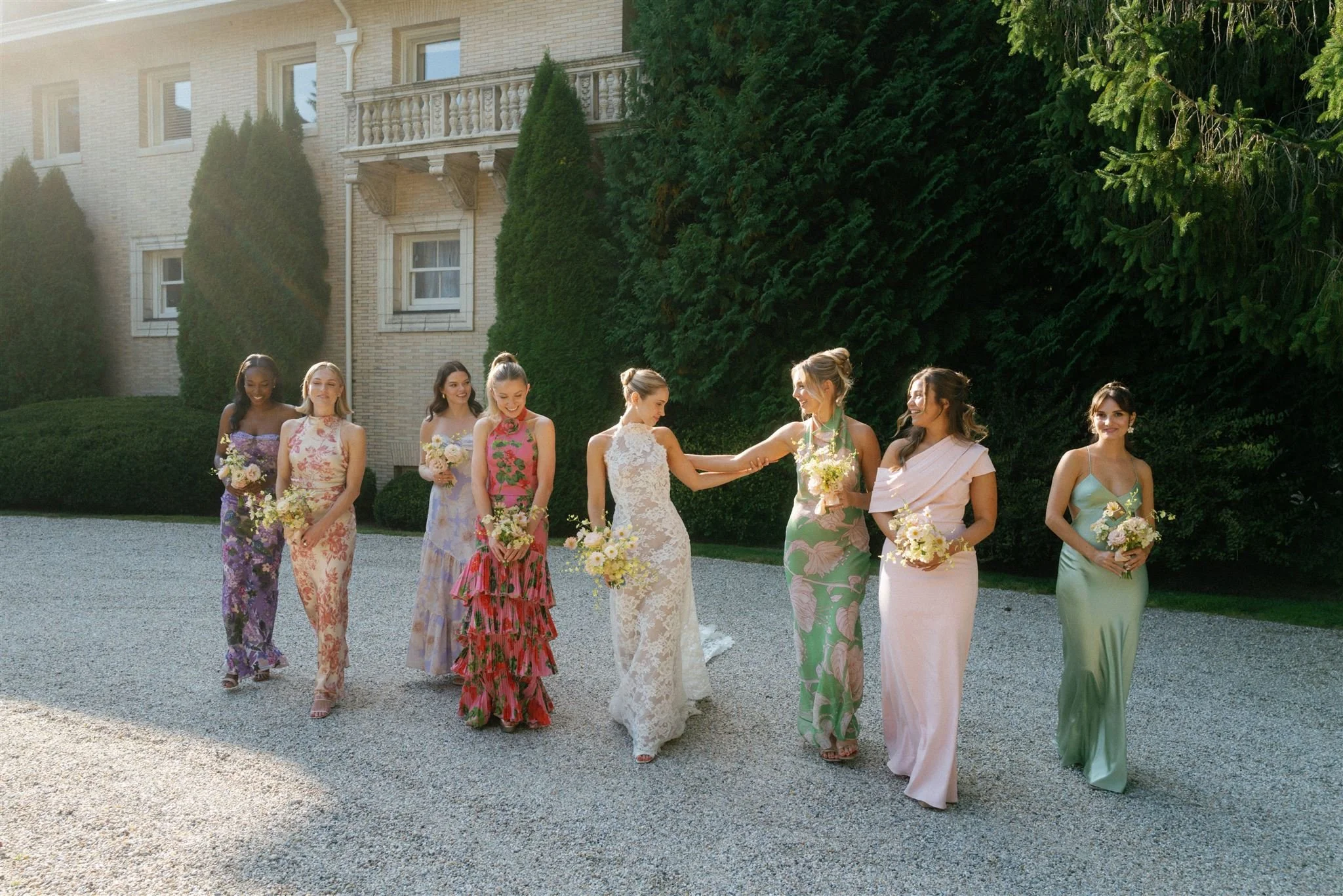 A bride in a white lace wedding dress and eight bridesmaids in colorful floral dresses walking outside on a gravel path.