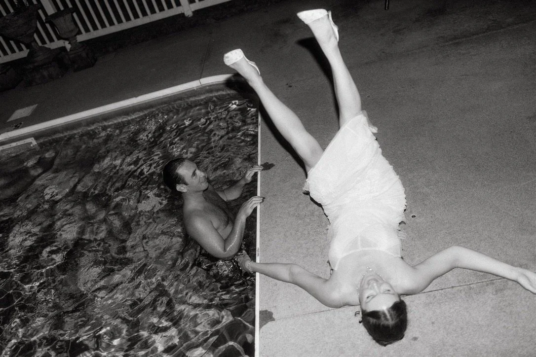 A woman in a white dress lying on the ground next to a swimming pool, smiling, with her head toward the camera and her legs raised in the air, while another woman in the pool holds a measuring stick.
