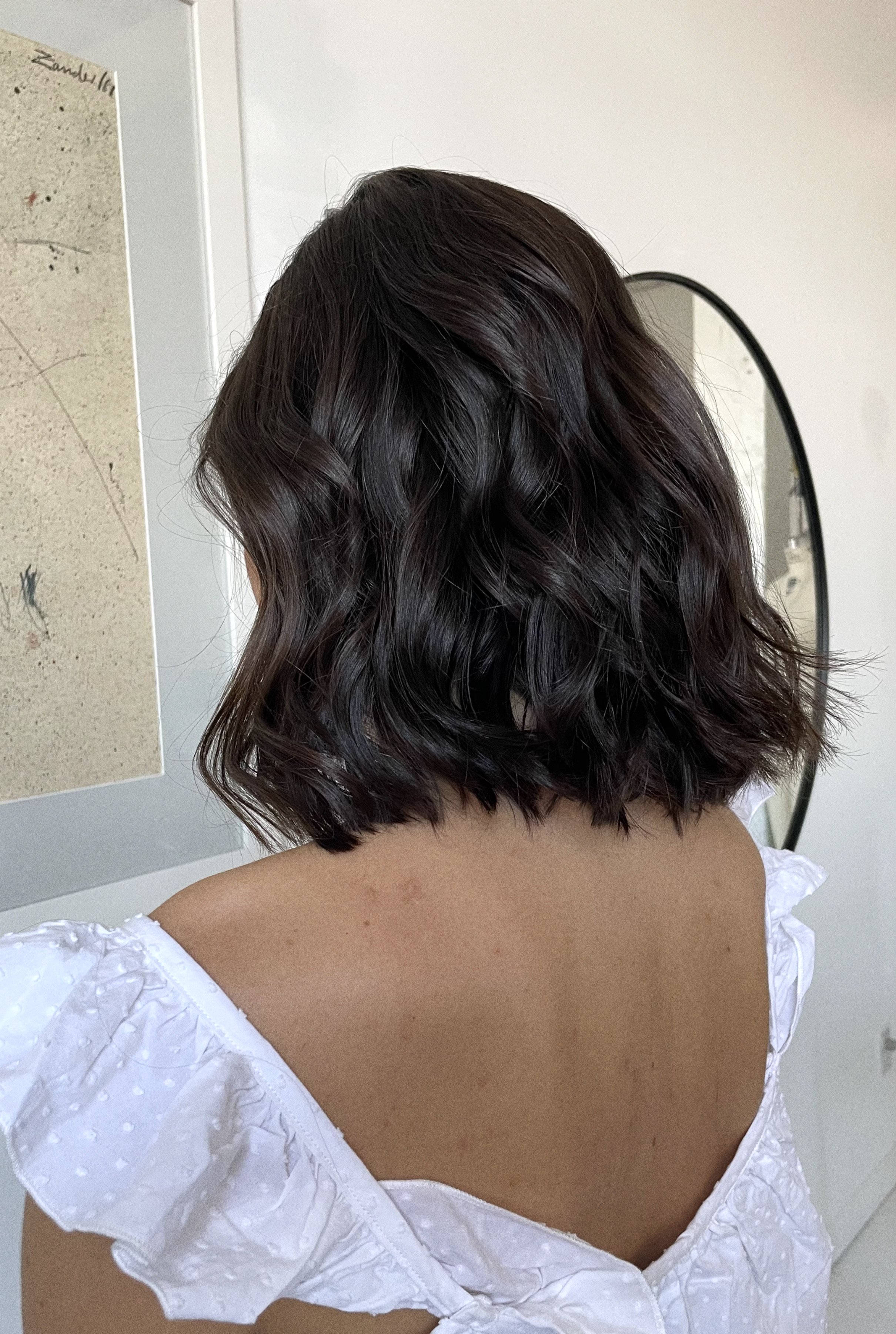 Back of a woman's head with shoulder-length wavy dark hair, wearing a white top with puffed sleeves, in front of a mirror and a white wall.