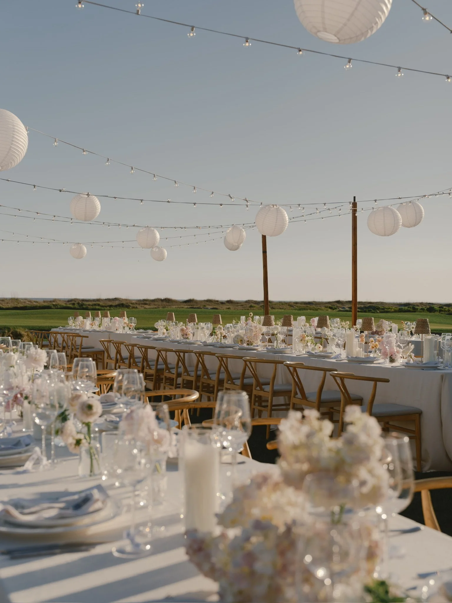 Outdoor banquet setup with long tables decorated with flowers, glassware, and candles, under hanging paper lanterns and string lights at sunset.