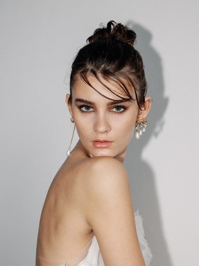 A young woman with styled up hair, wearing earrings, looking directly at the camera against a plain wall.
