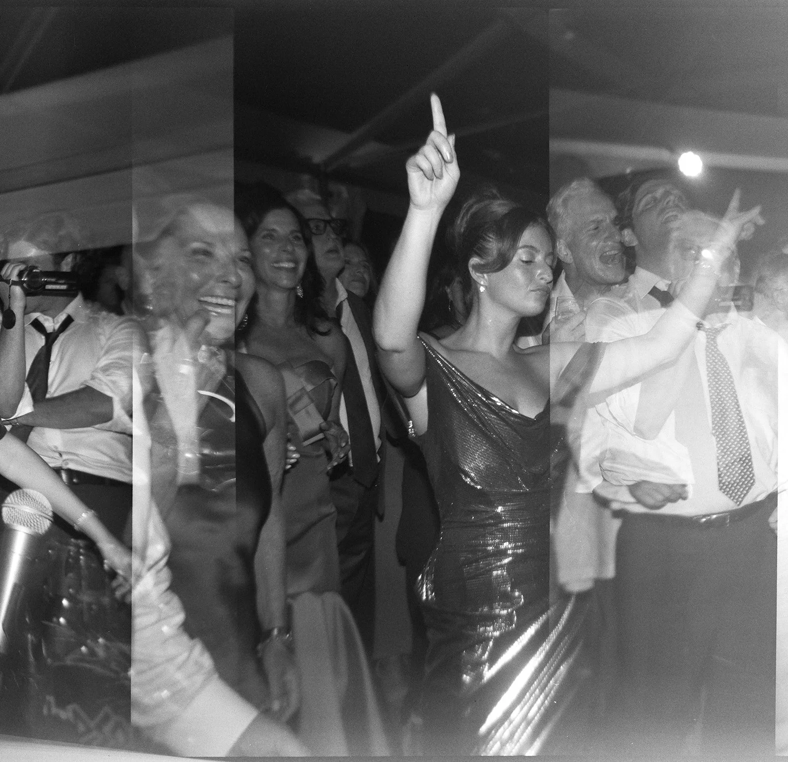 Black and white photo of a group of people at a celebratory event. Women and men are smiling, laughing, and making gestures, with one woman in a shimmering dress raising her hand. The setting appears festive and lively.