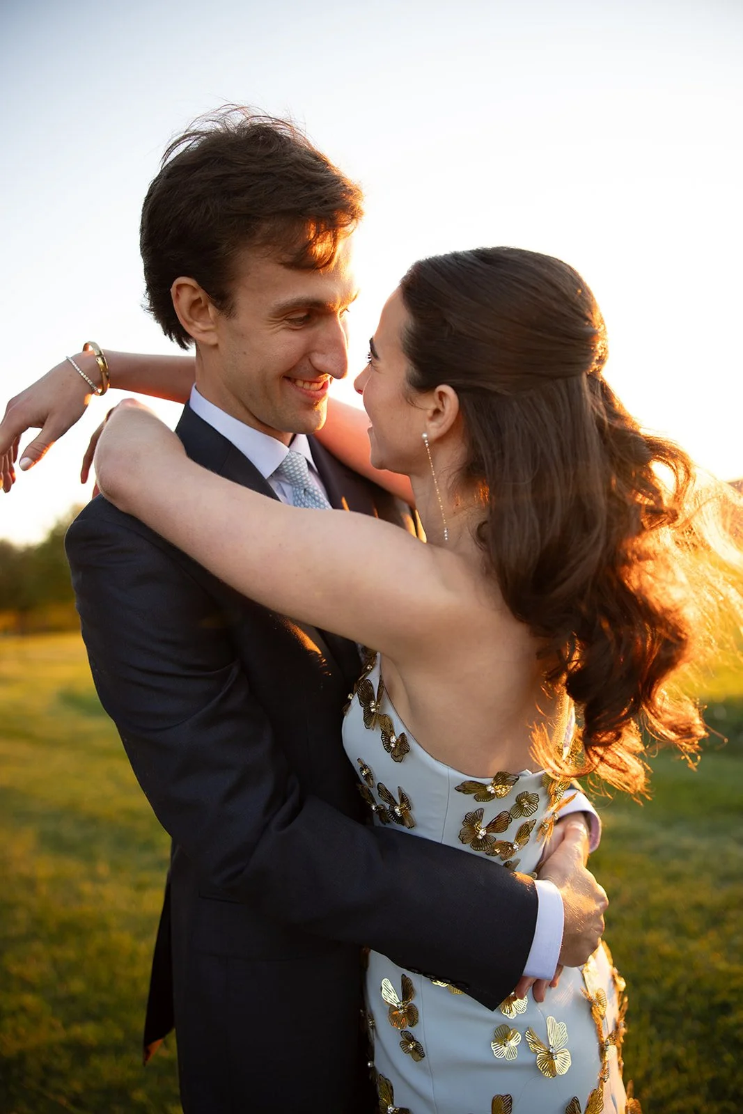 A young couple in formal attire sharing a romantic embrace outdoors at sunset.