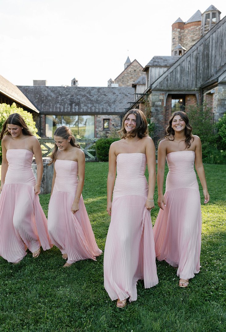 Four women in matching pink strapless dresses walking on a grassy lawn with a stone building in the background.