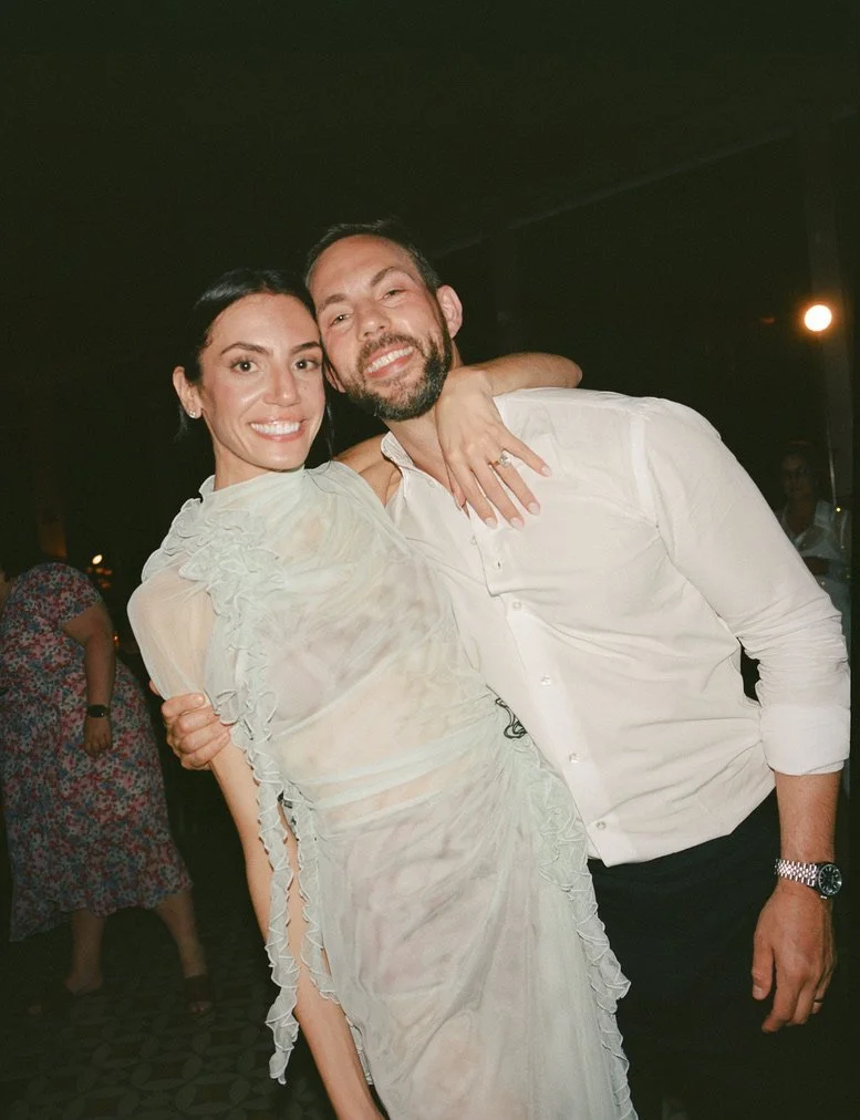 A man and woman smiling at the camera, with the woman giving the man a hug. The woman has dark hair and is wearing a light-colored, ruffled dress. The man has a beard, dark hair, and is dressed in a white shirt with rolled-up sleeves. They are indoor