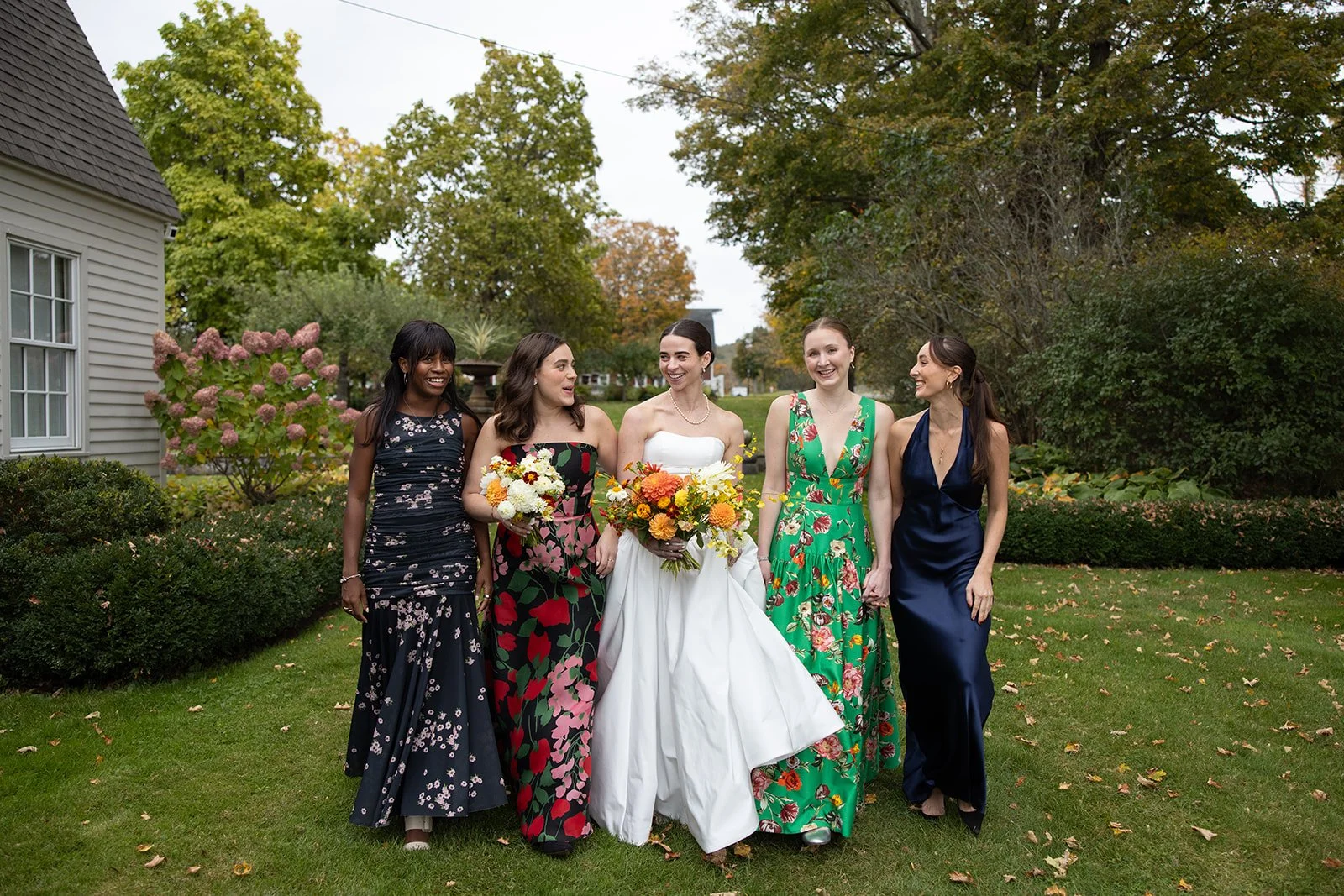 Group of women dressed in formal attire walking outdoors in a garden with trees and bushes, one woman in a white wedding dress holding a bouquet, others holding flowers, all smiling and talking.